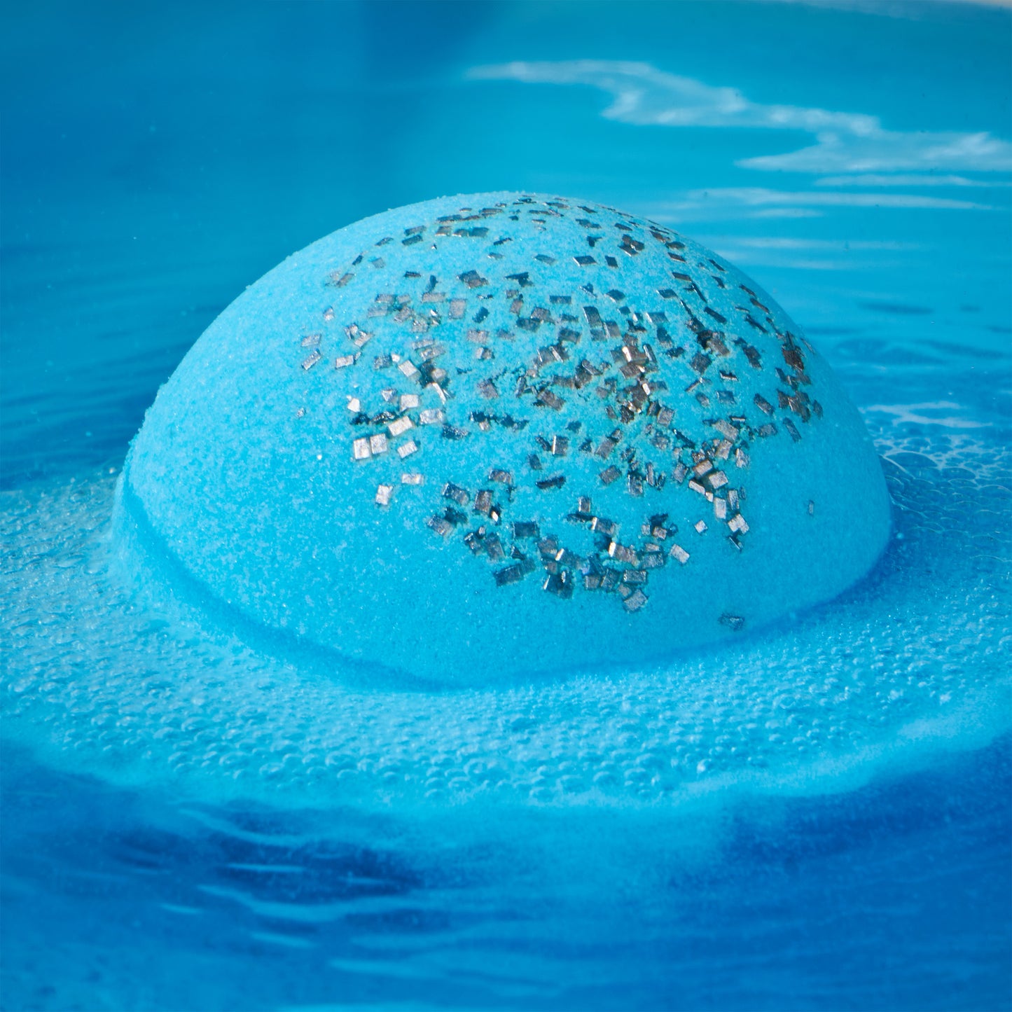 a blue bath bomb with sprinkles half submerged fizzing in water