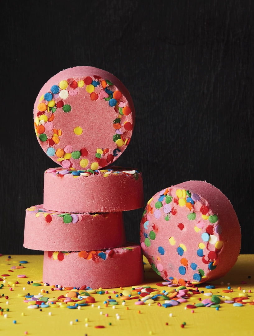 Stack of pink Candy Crush bath bombs topped with colorful candy sprinkles, set against a black background with sprinkles scattered on a yellow surface.