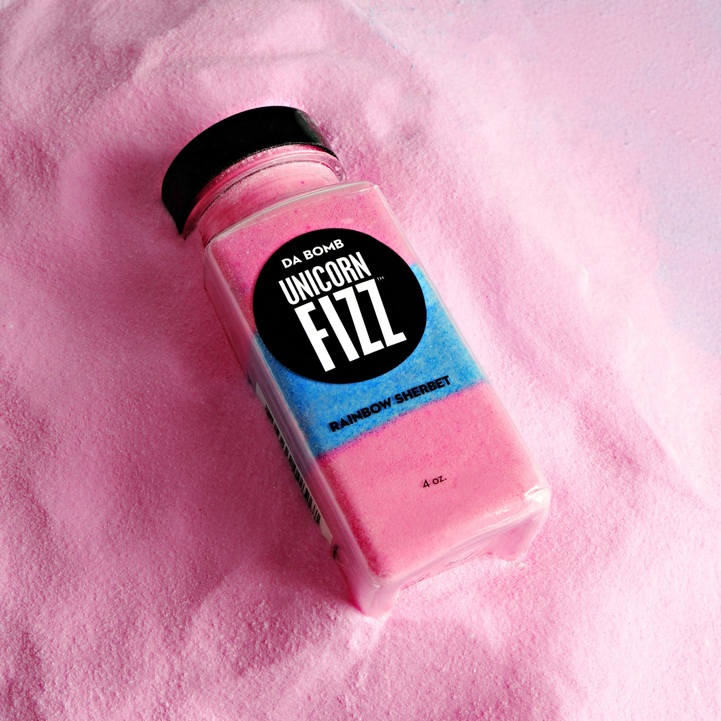 Da Bomb Unicorn Fizz in rainbow sherbet scent, resting on pink fizzy powder with layered pink and blue colors inside a clear 4 oz. container.