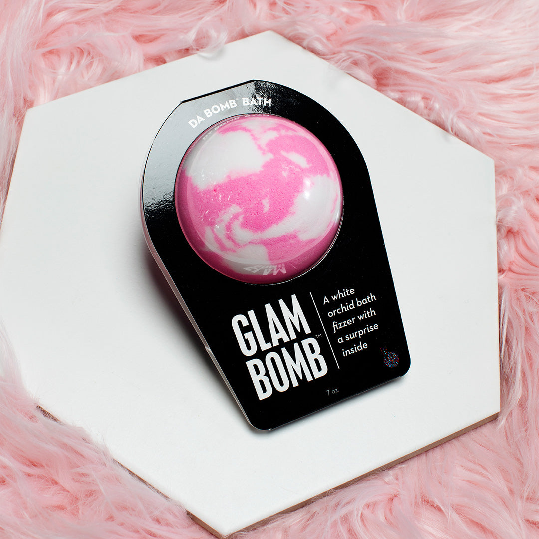 Pink bath bomb with white swirls in black packaging sitting on a white hexagram plate surrounded by pink fuzz