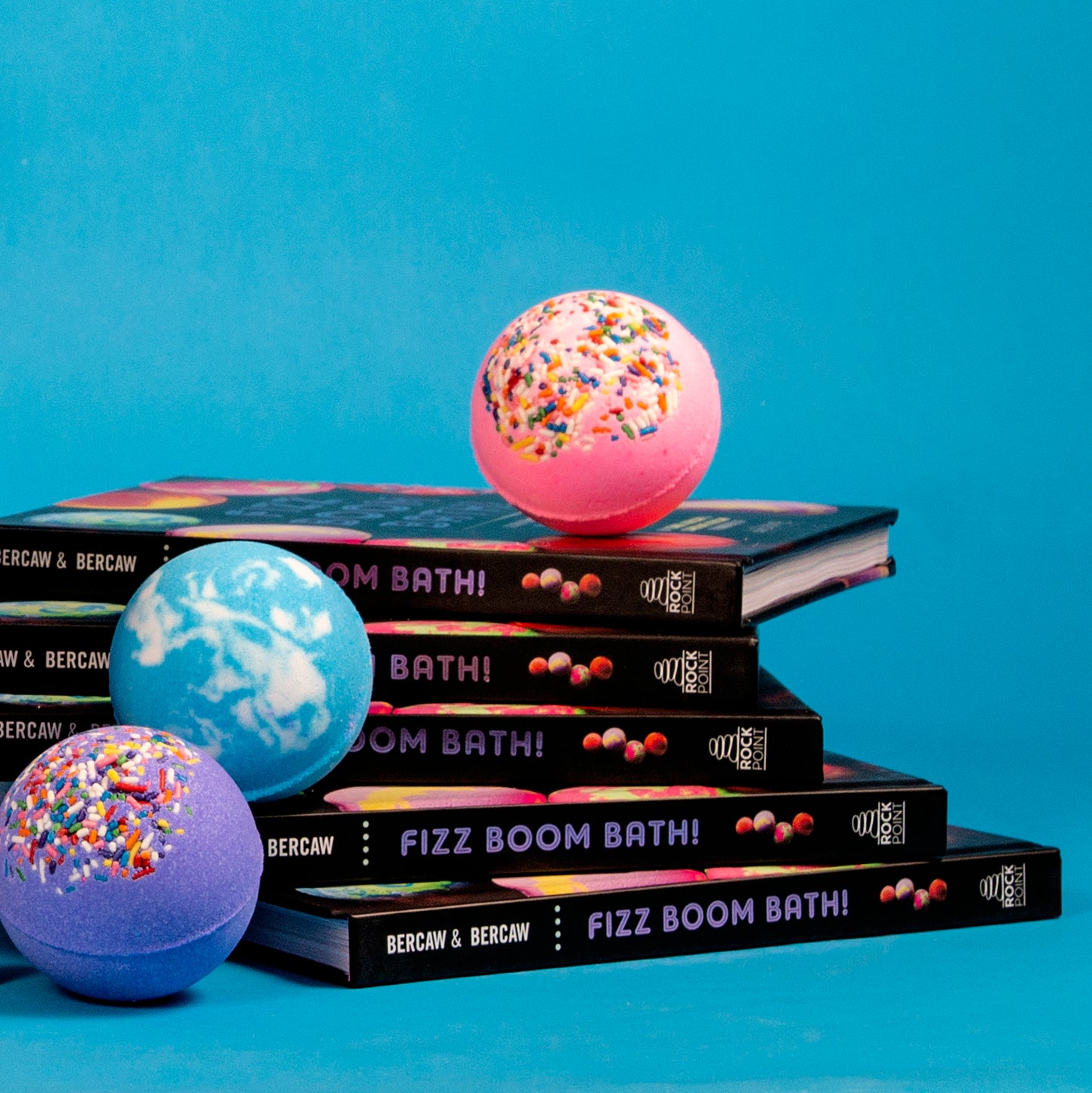 DIY bath bomb books titled Fizz Boom Bath! stacked with colorful Da Bomb bath bombs on top, including sprinkle-covered and swirled designs, set against a bright blue background.