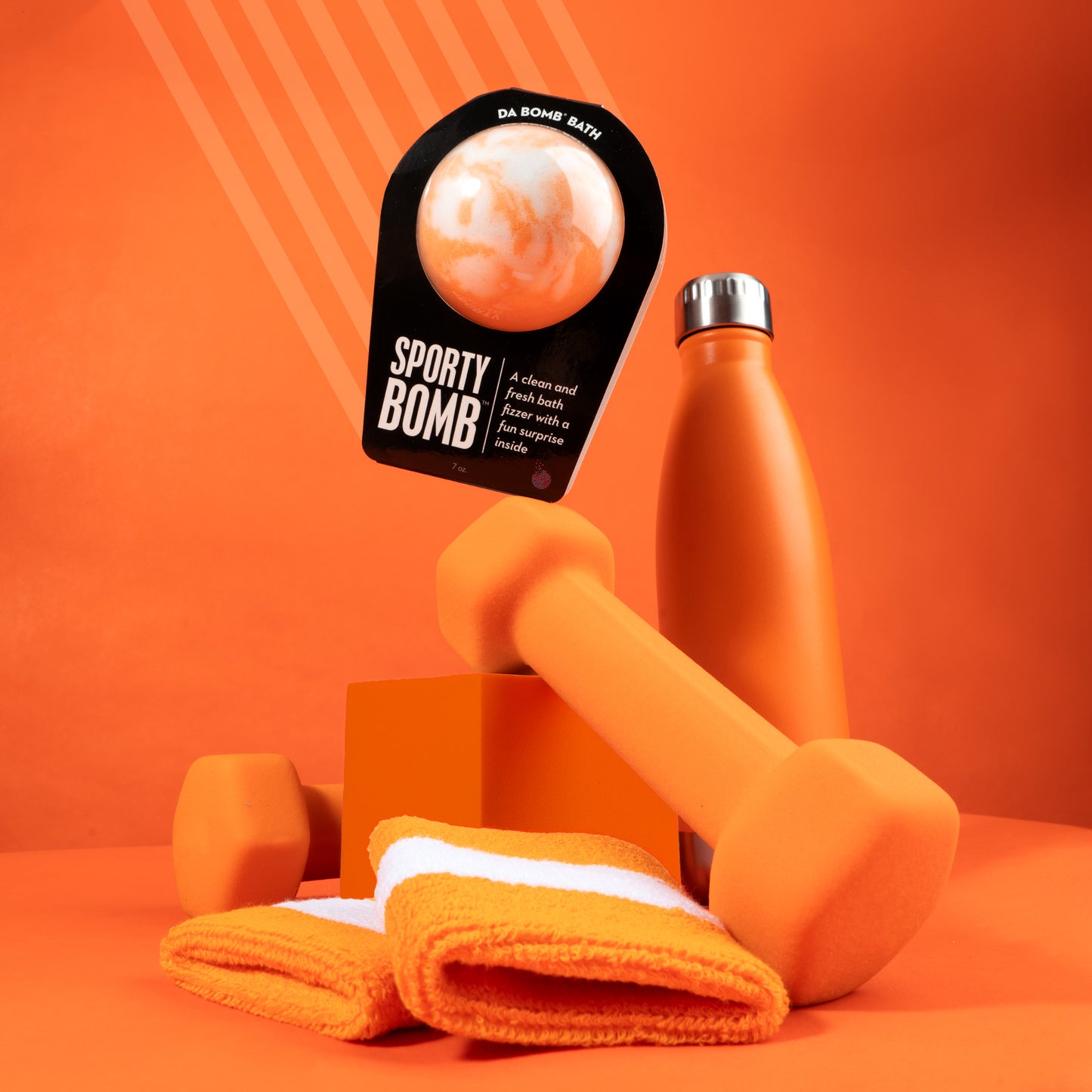 Orange bath bomb in black packaging balance don an orange dumbbell. Surrounded by an orange water bottle, sweatbands, and weights, on an orange background