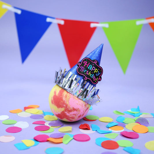 bath bomb wearing a party hat with confetti on ground and bunting in background on light purple background