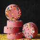 Stack of pink Candy Crush bath bombs topped with colorful candy sprinkles, set against a black background with sprinkles scattered on a yellow surface.