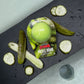 Green pickle bath bomb on black board over a tub filled with water surrounded by pickle slices and spears