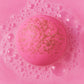 Mom Bomb bath fizzer dissolving in bright pink water with soft bubbles. 